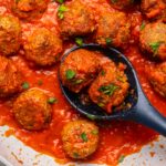 Vegan Meatballs in saute pan with marinara sauce topped with fresh parsley. A wooden spoon holds 2 meatballs in the pan