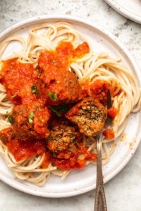 Vegan Meatballs tossed in marinara and served over spaghetti on white speckled plate. One meatball is cut in half to show the inside texture