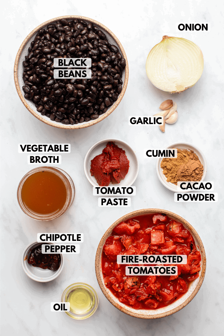 Ingredients for chili in small bowls on marble backrground. Clockwise text labels read onion, garlic, cumin, cacao powder, fire-roasted tomatoes, oil, chipotle pepper, tomato paste, vegetable broth, and black beans