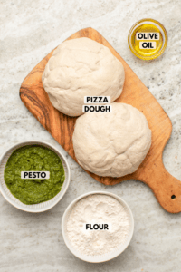Ingredients for pesto star bread on stone background. Clockwise text labels read olive oil, pizza dough, flour, and pesto