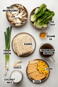 Ingredients for vegan ramen on stone countertop. Clockwise text labels read pak choy, sesame oil, mushroom broth, corn, tofu, coconut milk, garlic, green onion, ramen, and mixed mushrooms