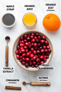 Ingredients for vegan cranberry sauce in small bowls on marble background. Clockwise text labels read maple syrup, orange juice, orange zest, fresh cranberries, nutmeg, cinnamon, and vanilla extract