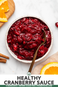 Bowl of cranberry sauce with gold serving spoon on wooden serving board surrounded by orange sliced and cinnamon sticks