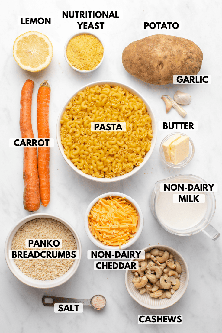 Ingredients for vegan mac and cheese in small white bowls arranged on a marble background. Clockwise text labels read nutritional yeast, potato, garlic, butter, non-dairy milk, cashews, non-dairy cheddar, salt, panko breadcrumbs, carrot, and lemon.