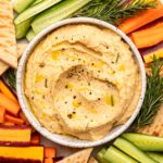 Small white bowl of butternut squash hummus on veggie tray with pita, carrots, and cucumber