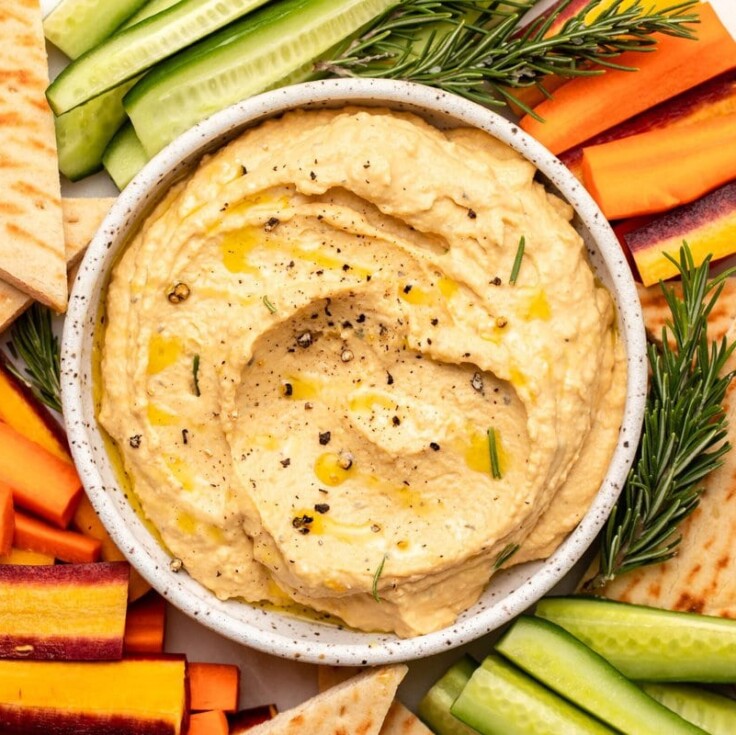 Small white bowl of butternut squash hummus on veggie tray with pita, carrots, and cucumber