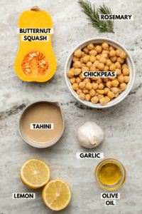 Ingredients for hummus on stone countertop. Clockwise text labels read rosemary, chickpeas, garlic, olive oil, lemon, tahini, and butternut squash
