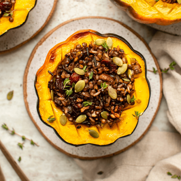 Stuffed acorn squash topped with pumpkin seeds on small white plate, surrounded by other cooked squash on plates
