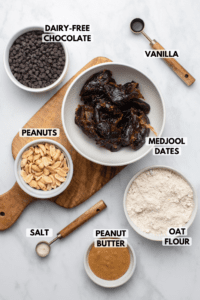 Ingredients for Vegan Snickers bars in small bowls on marble background. Clockwise text labels read vanilla, medjool dates, oat flour, peanut butter, salst, peanuts, and dairy-free chocolate
