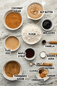 Ingredients for ginger molasses cookies on stone countertop. Clockwise text labels read nut butter, molasses, flour, vanilla, baking soda, salt, nutmeg, ginger, cinnamon, cloves, turbinado sugar, maple syrup, flax egg, and coconut sugar