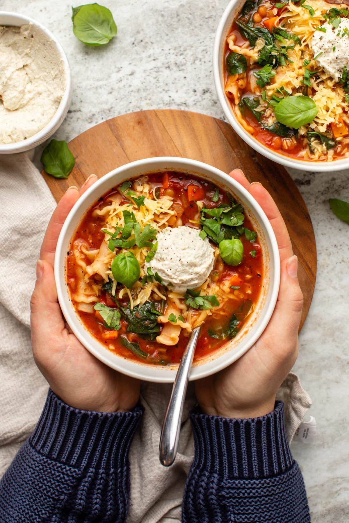 Hands holding a bowl of vegan lasagna soup topped with vegan ricotta cheese and fresh basil