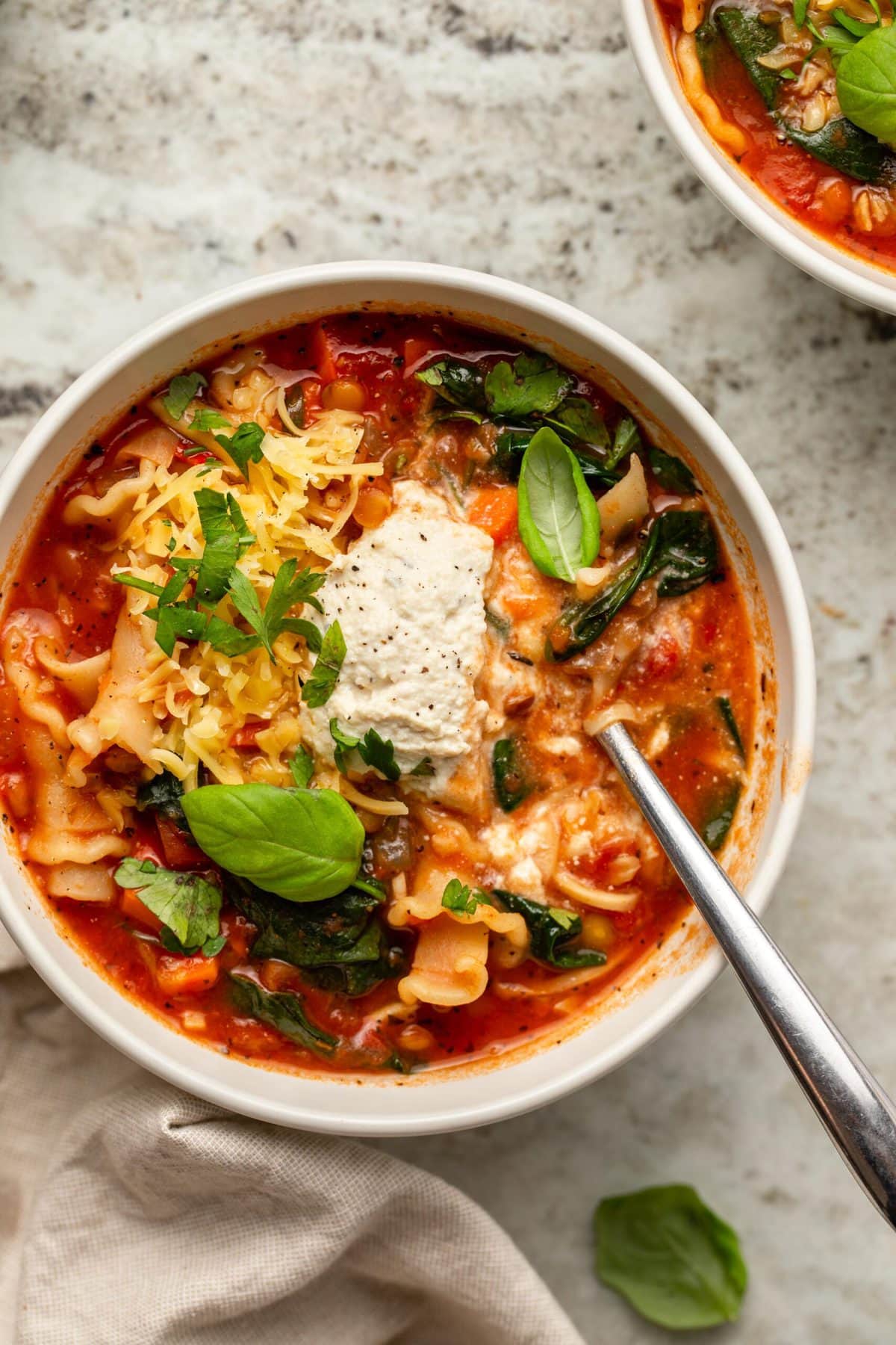Close-up photo of vegan lasagna soup topped with ricotta cheese, and fresh herbs on stone countertop