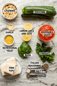 Ingredients for Vegan Zucchini Rolls arranged on kitchen countertop. Clockwise text labels read cashews, zucchini, marinara sauce, parsley, italian seasoning, salt, pepper, firm tofu, basil, nutritional yeast, lemon juice and zest, and garlic