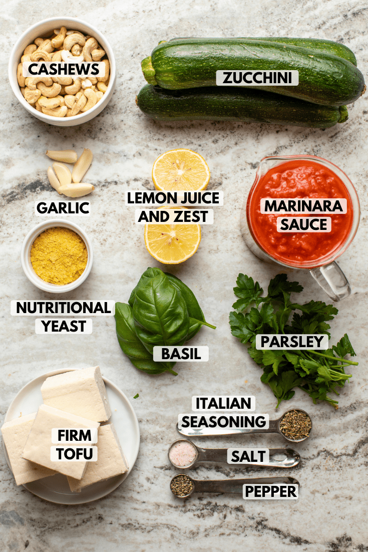 Ingredients for Vegan Zucchini Rolls arranged on kitchen countertop. Clockwise text labels read cashews, zucchini, marinara sauce, parsley, italian seasoning, salt, pepper, firm tofu, basil, nutritional yeast, lemon juice and zest, and garlic