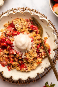 Strawberry rhubarb crisp in white dish topped with melty vanilla ice cream