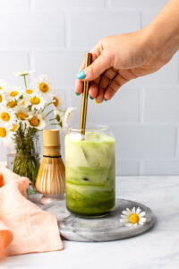 hand stirring plant milk into matcha and chamomile drink in glass with ice