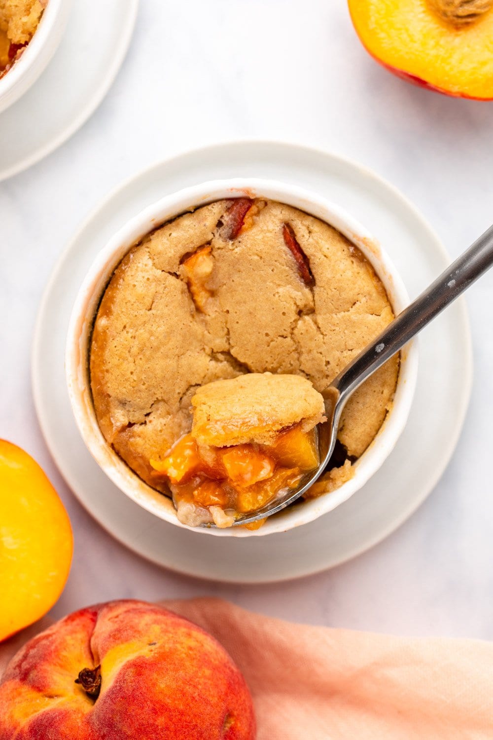 peach cobbler with spoon taking scoop out of it on white marble background