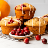 three vegan cranberry orange muffins stacked on top of a baking tray with several cranberries spread around