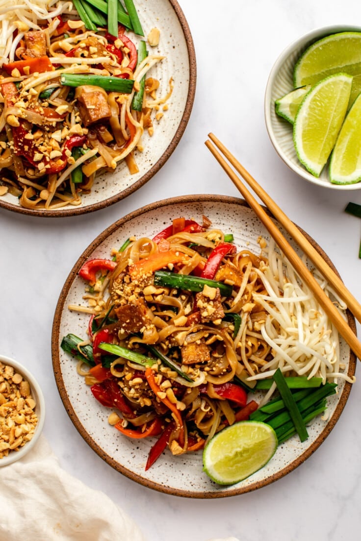 two plates of pad thai with lime wedges and crushed peanuts