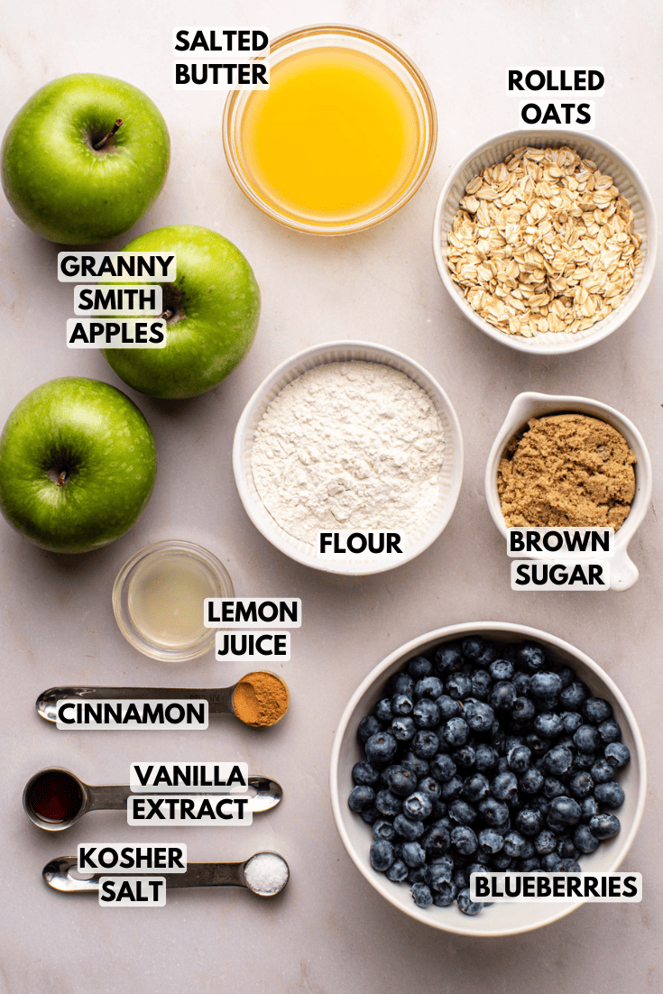 ingredients for apple blueberry crisp laid out on a marble kitchen countertop served in various sizes of white bowls