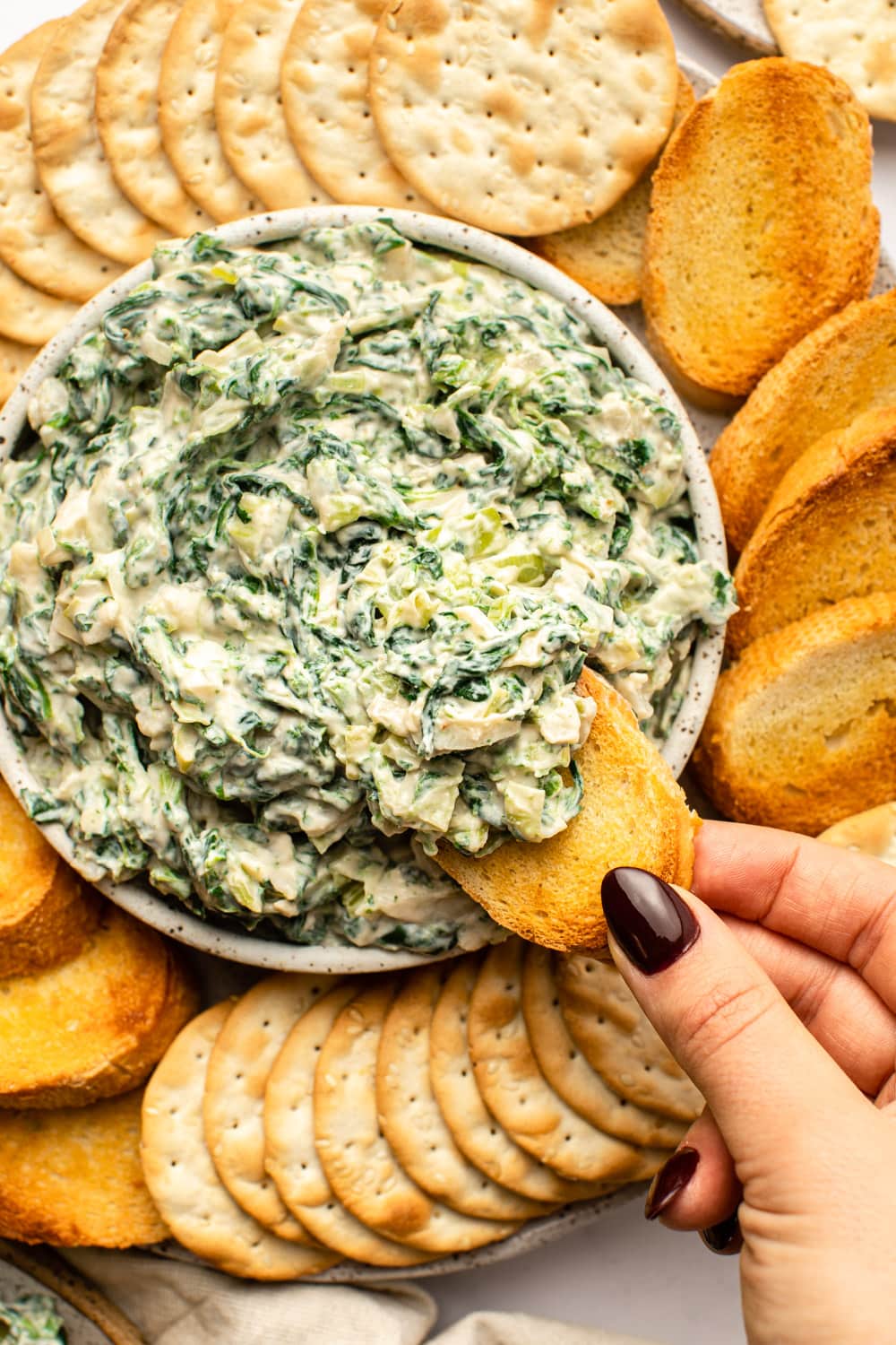 vegan spinach dip served in a white bowl with crackers and toasted crostini