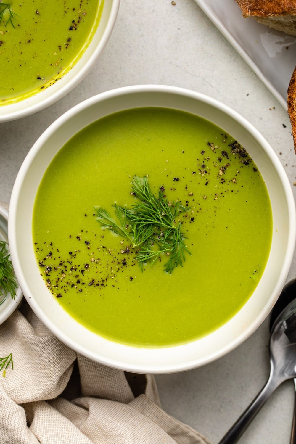 green pea soup served in a white bowl and topped with fresh dill and black pepper with another bowl of soup and fresh bread laid on a marble kitchen countertop