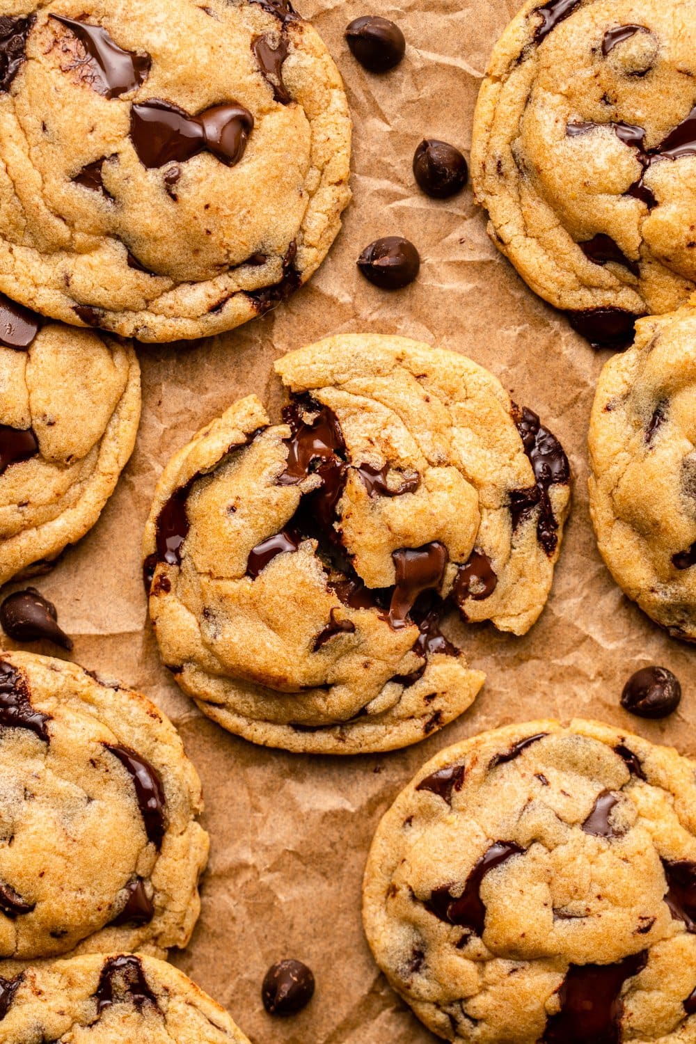 seven chocolate chip cookies laid out on parchment paper with the cookie in the middle divided into two to showcase the melting chocolate