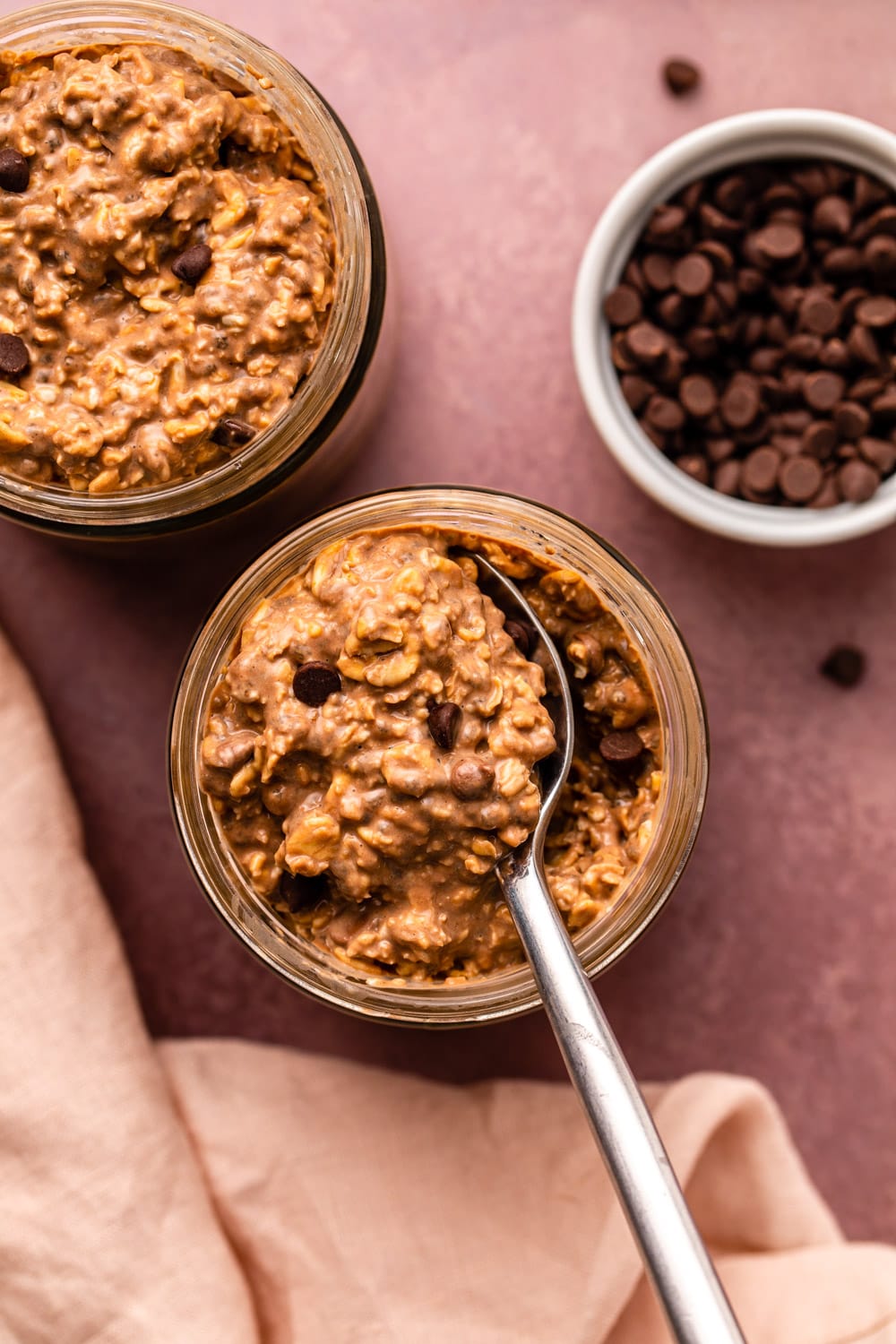 two jars of chocolate overnight oats and a chocolate chips in a small white jar placed on a burgundy kitchen countertop