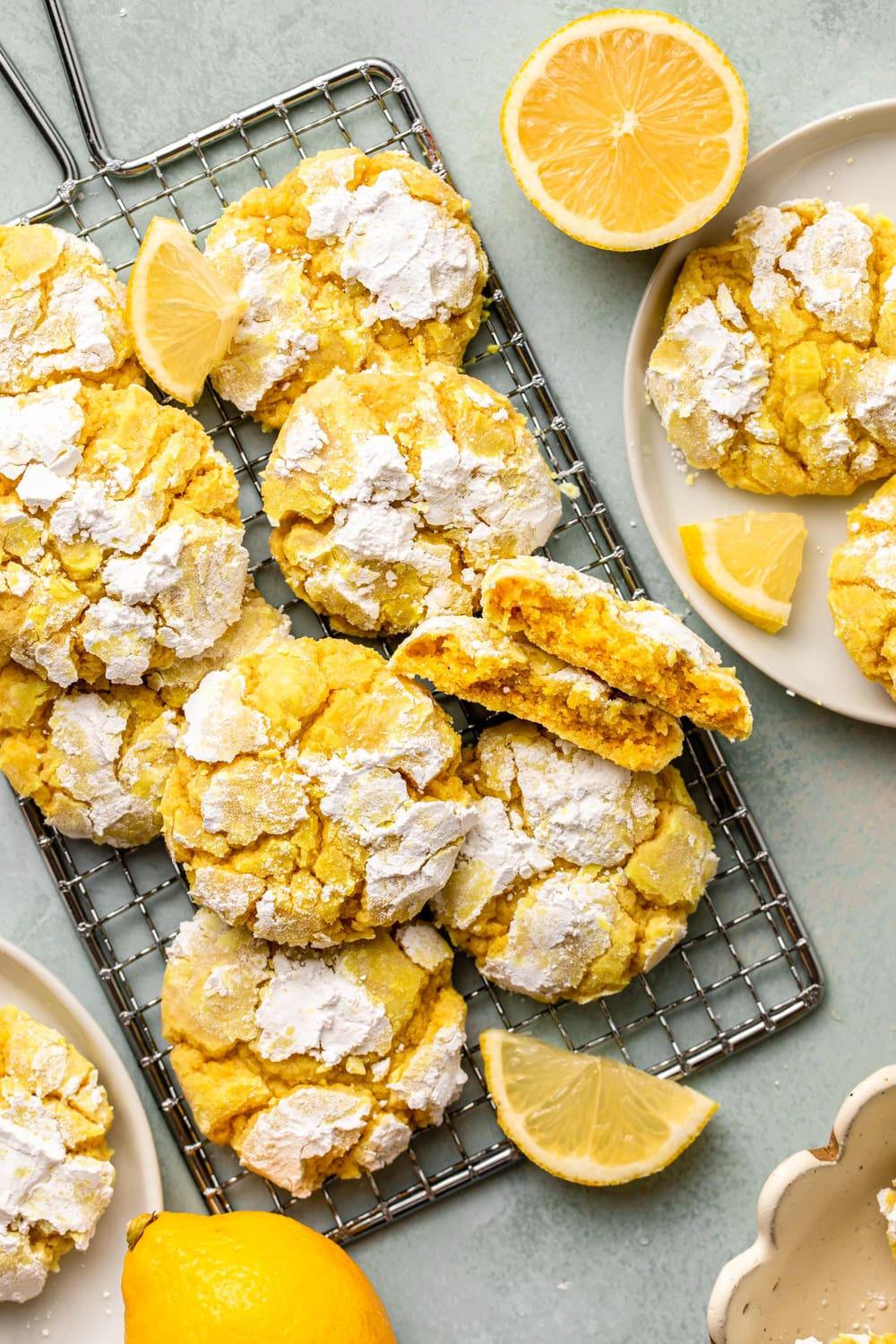 lemon crinkle cookies cooling on a cooling rack next to a plate of lemon cookies and half of a lemon, on a sea green kitchen countertop