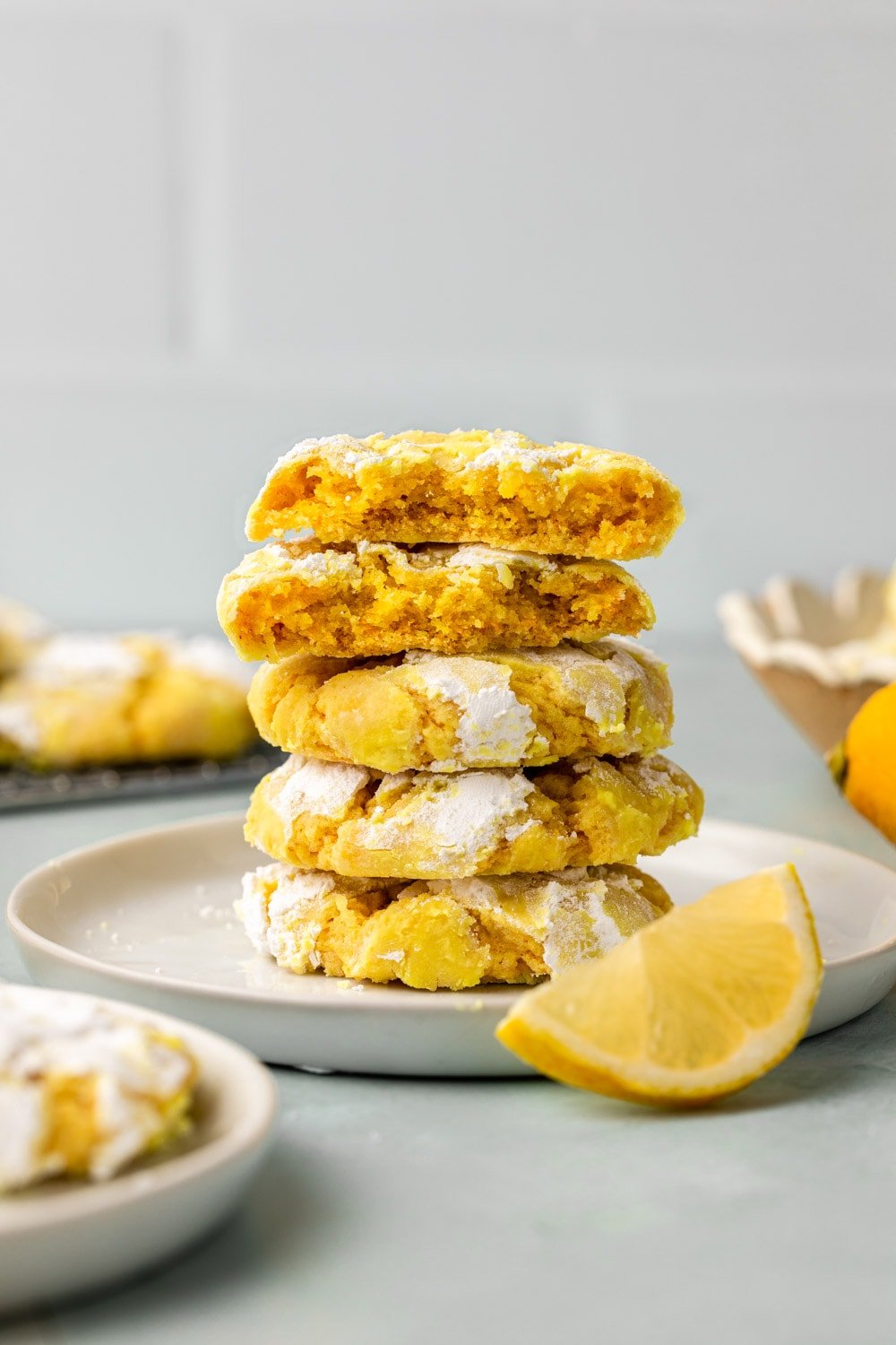 four lemon crinkle cookies stacked on top of each other on a white plate with a slice of lemon placed next to the plate, in a white backdrop