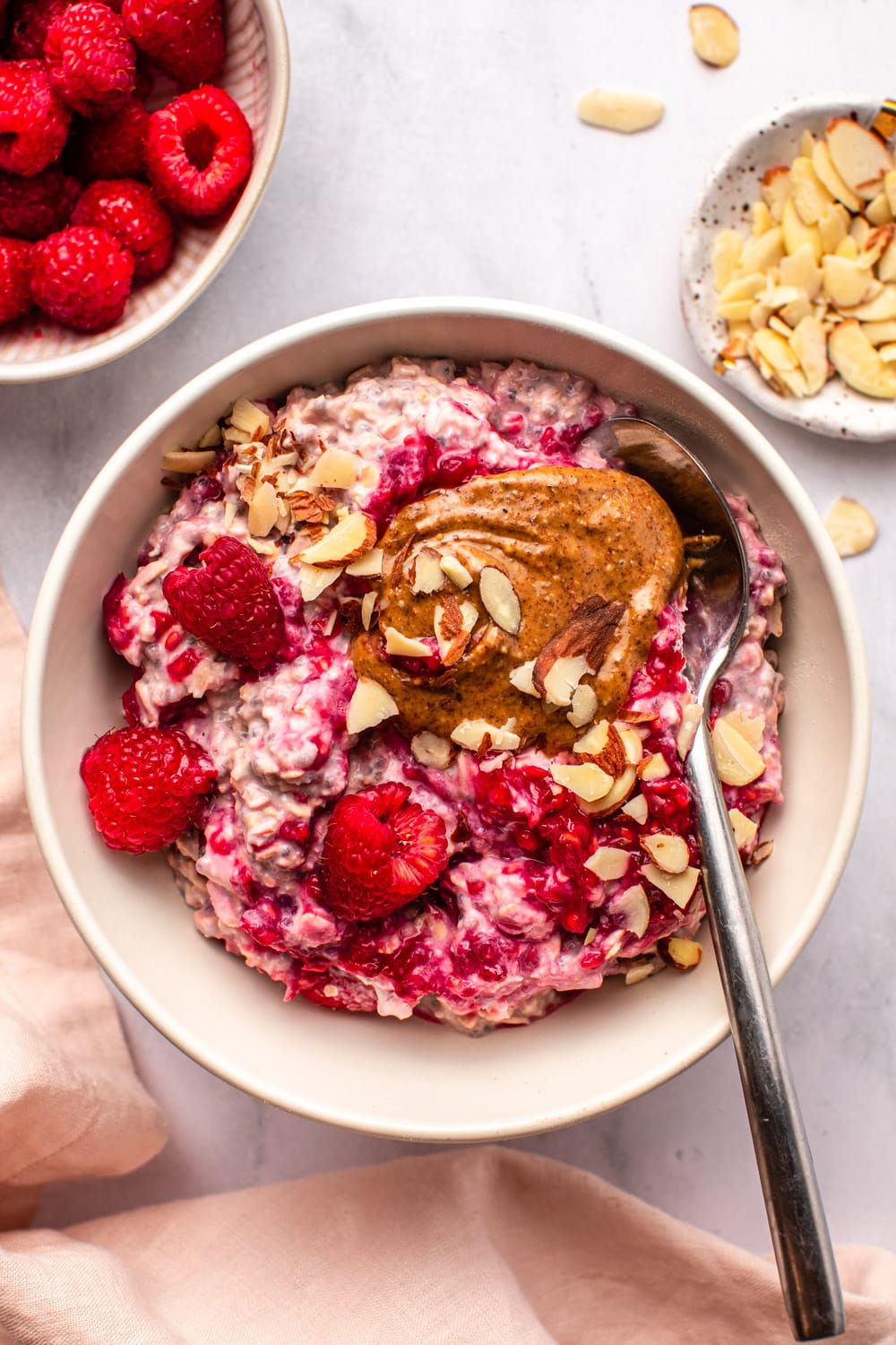 raspberry overnight oats served on a white bowl topped with fresh raspberries, crushed almonds, almond butter and a spoon