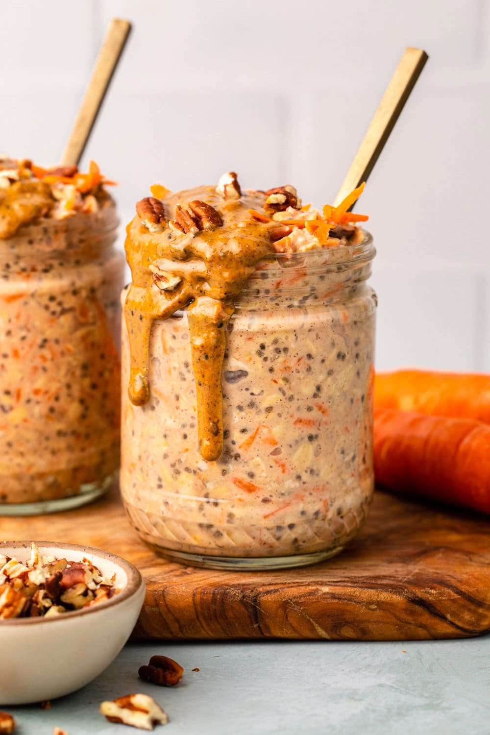 a jar of carrot cake overnight oats placed on a cutting board with a small bowl of chopped pecans and another jar of carrot cake overnight oats and two carrots set aside
