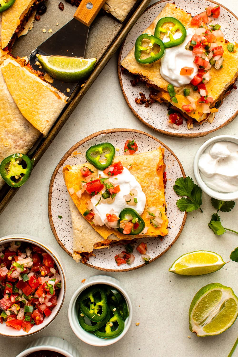 a slice of sheet pan quesadillas served on a plate topped with salsa, sour cream and two slices of jalapeno, placed on a marble kitchen countertop surrounded by the tray of quesadillas and several bowls served with salsa, sliced jalapenos, sour cream and slices of lime
