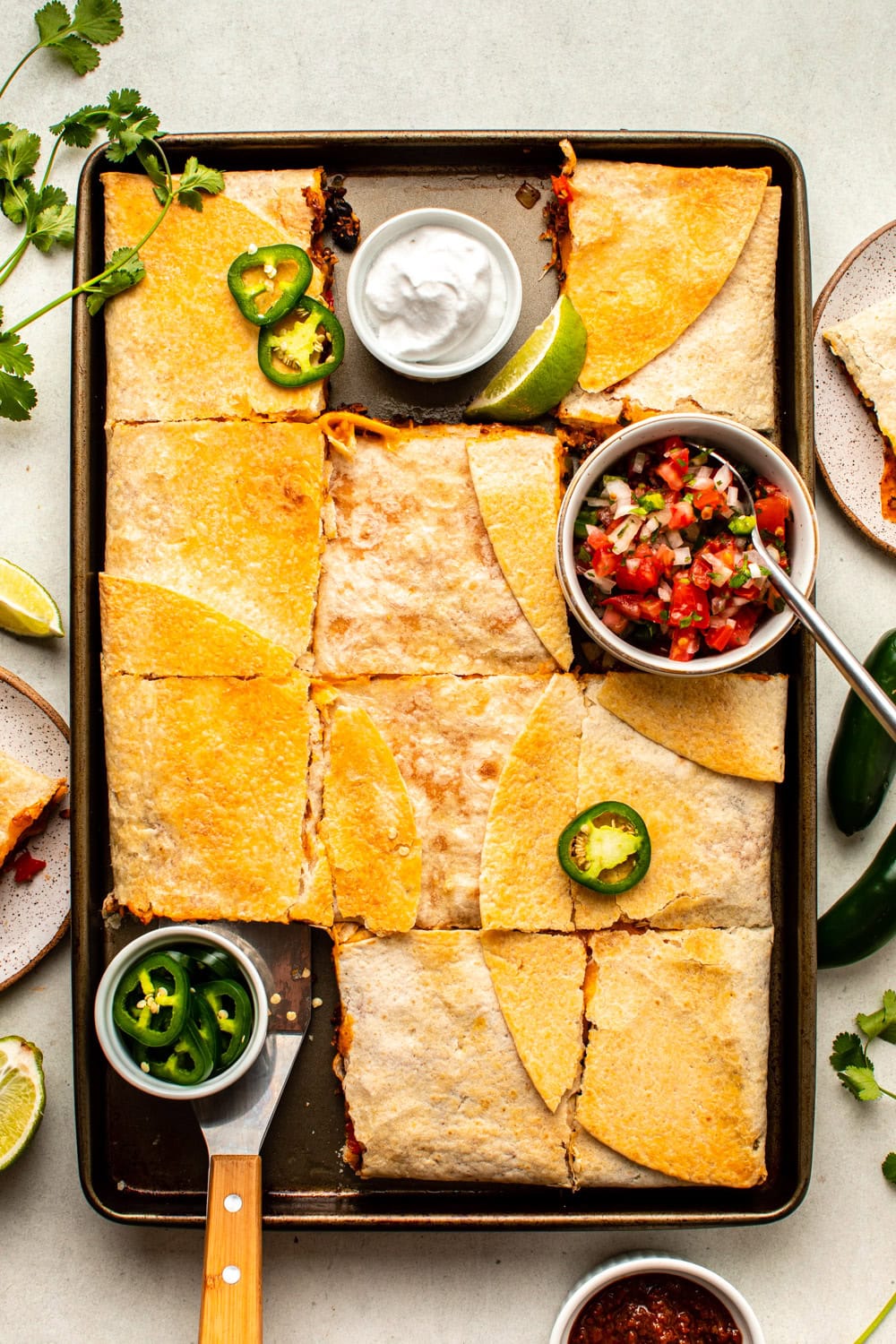 a photo of the baked quesadilla served in a baking tray topped with small bowls of salsa and sliced jalapenos placed on the empty corners of the tray