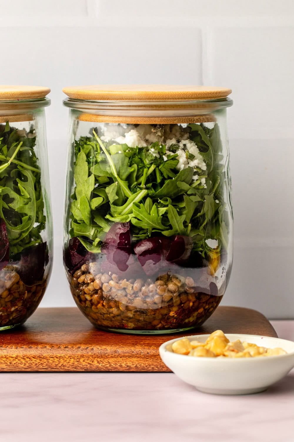 two jars of beet and lentil salad placed on a cutting board on a kitchen countertop, with a small bowl of hazelnuts in front as a topping ingredient