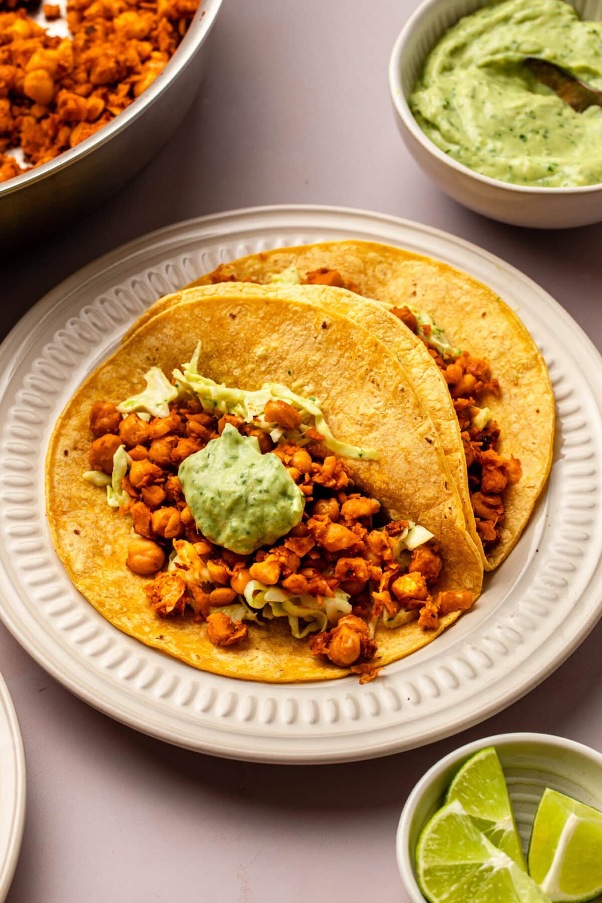 chickpea tacos served on a ceramic plate, with a small bowl of lime wedges, a side of avocado crema, and a baking tray visible in the background