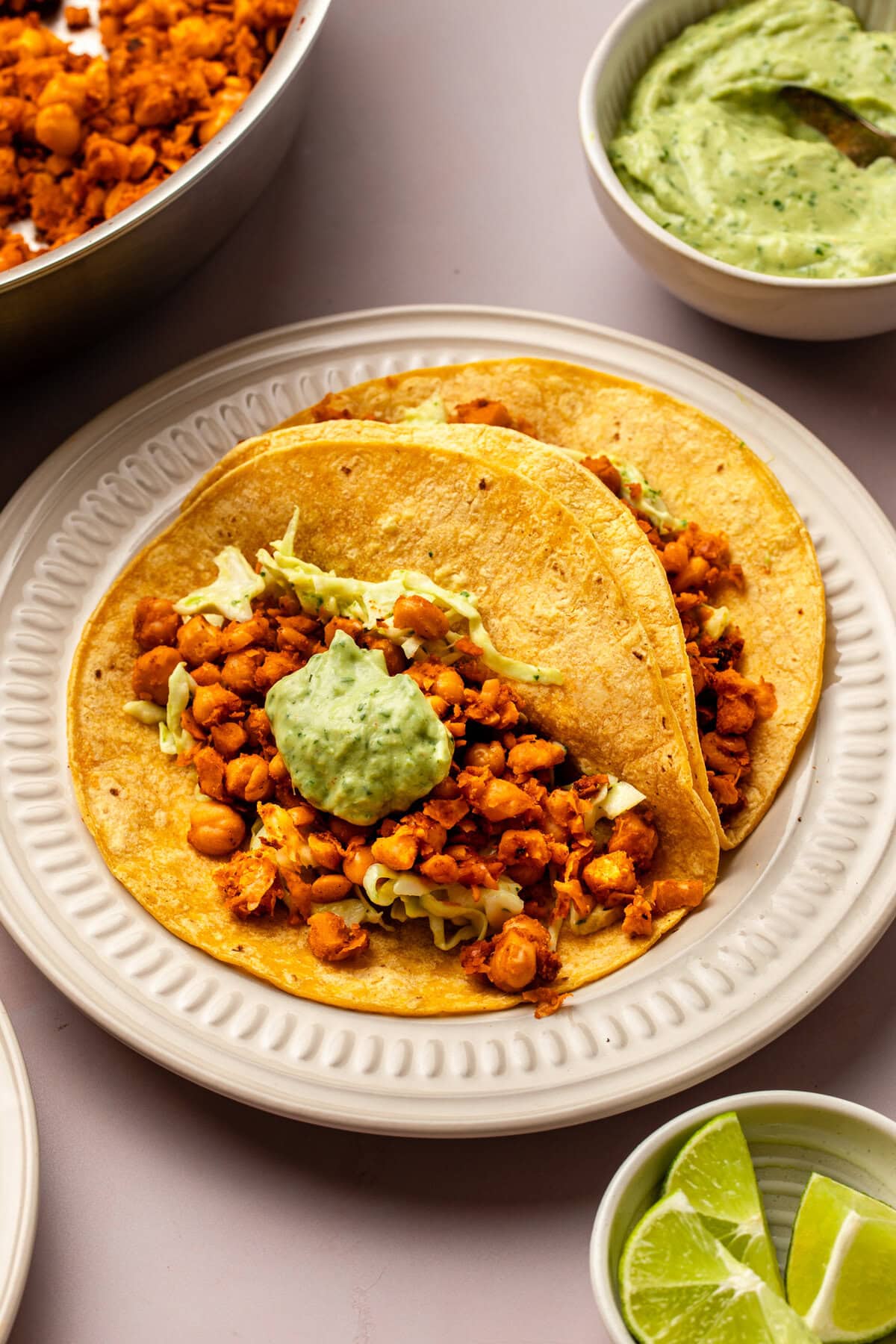 chickpea tacos served on a ceramic plate, with a small bowl of lime wedges, a side of avocado crema, and a baking tray visible in the background