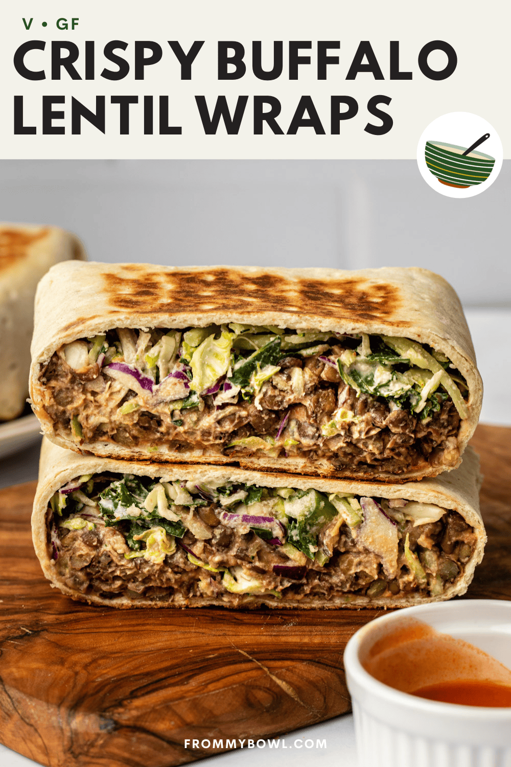 crispy lentil buffalo wrap cut in half on a wooden cutting board placed on a kitchen counter. A small bowl of buffalo sauce sits nearby.