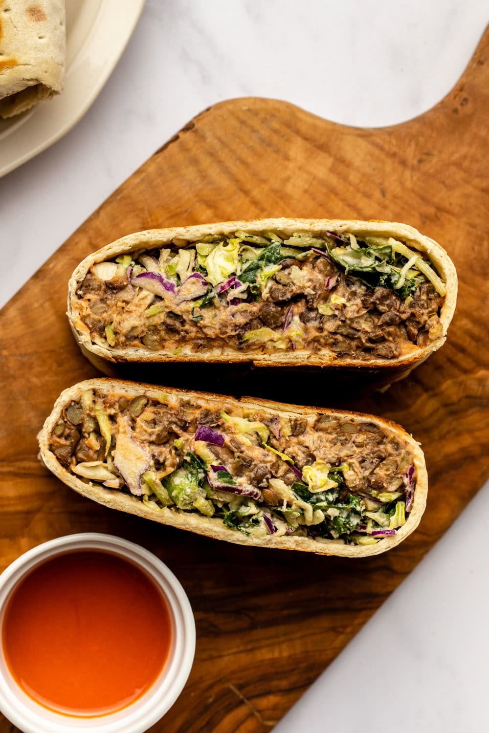 crispy lentil buffalo wrap cut in half on a wooden cutting board placed on a kitchen counter. A small bowl of buffalo sauce sits nearby.
