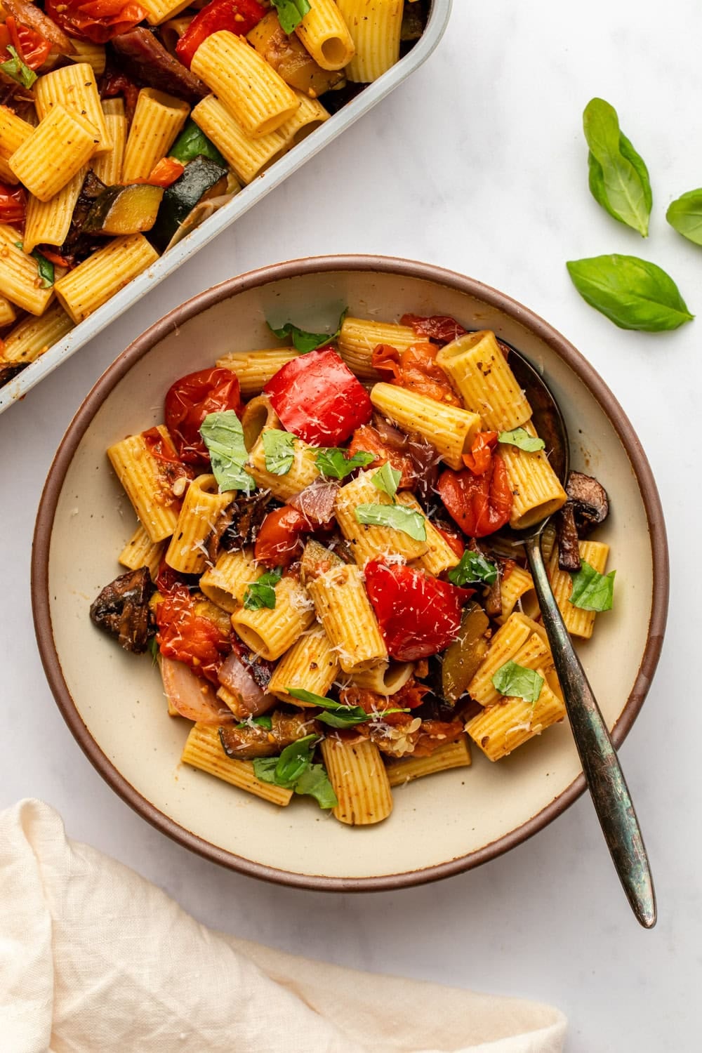 a ceramic plate of roasted vegetable pasta is served on a kitchen counter, with a spoon resting inside the dish. nearby, there&rsquo;s extra pasta, a cloth, and fresh basil leaves arranged around the scene.