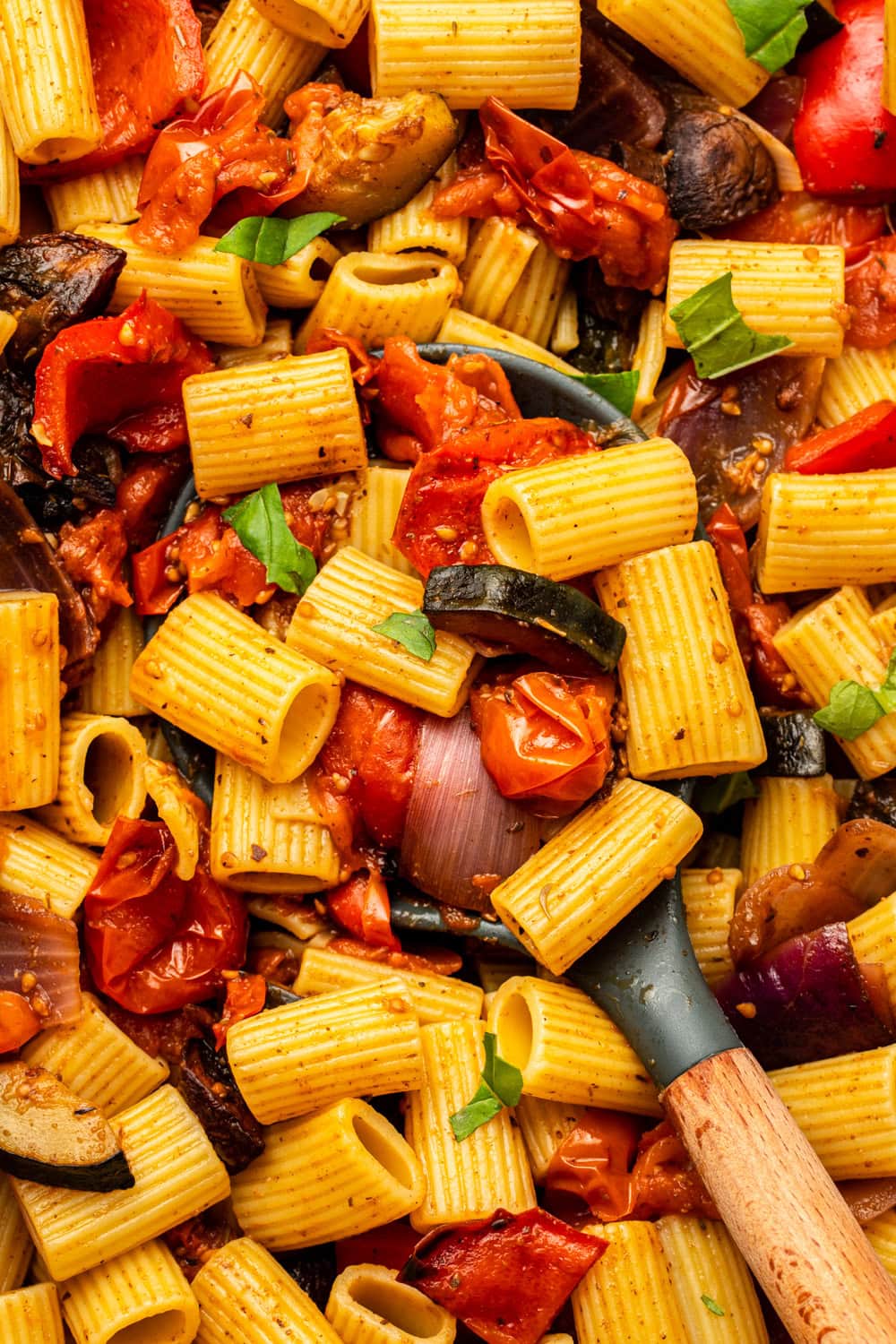 a close-up, zoomed-in shot of the roasted vegetable pasta, with a large serving spoon inside the dish as if stirring it.