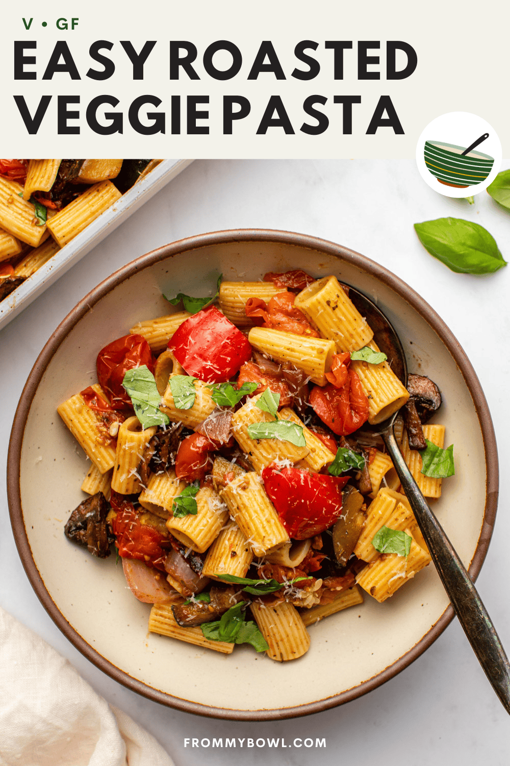a ceramic plate of roasted vegetable pasta is served on a kitchen counter, with a spoon resting inside the dish. nearby, there&rsquo;s extra pasta, a cloth, and fresh basil leaves arranged around the scene.