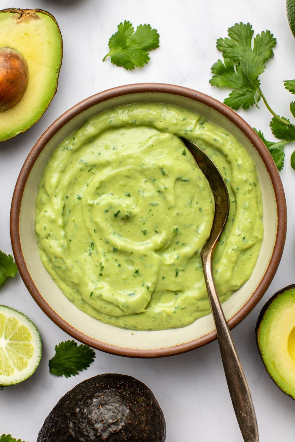 a ceramic bowl filled with vegan avocado crema sits on a kitchen countertop, with a serving spoon resting inside. around the bowl, fresh avocado halves, cilantro, and lime are arranged decoratively.