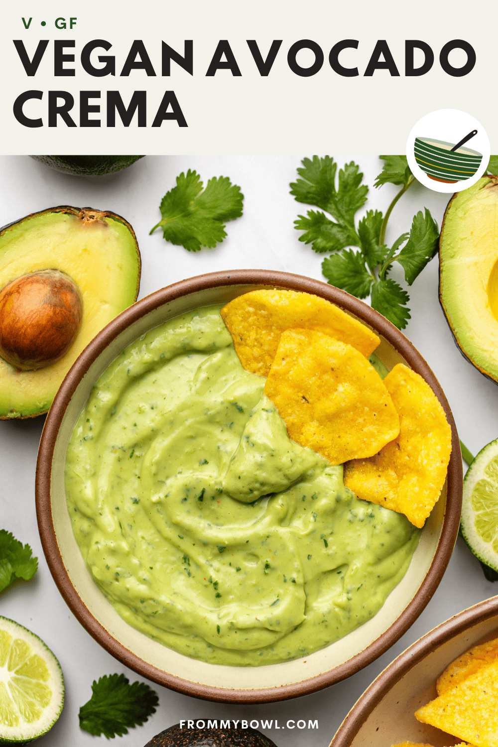 a ceramic bowl of vegan avocado crema sits on the countertop, topped with tortilla chips, with avocado, cilantro, and lime arranged decoratively around it.