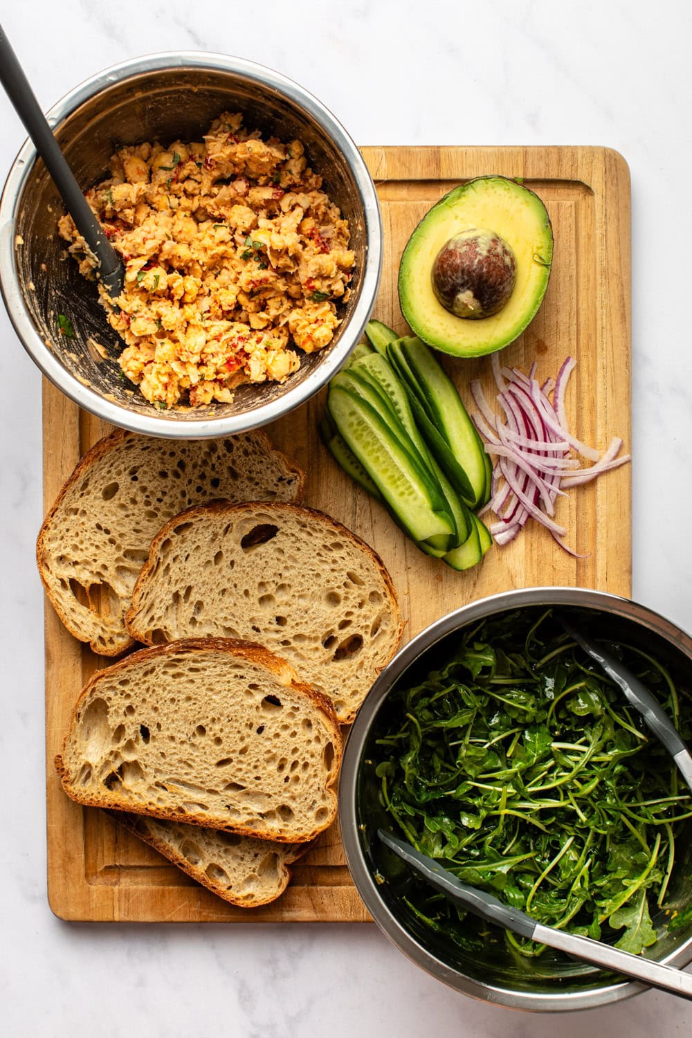 ingredients prepared for assembling smashed chickpea salad sandwiches, including sourdough bread, avocado, cucumber, arugula, and the chickpea filling.