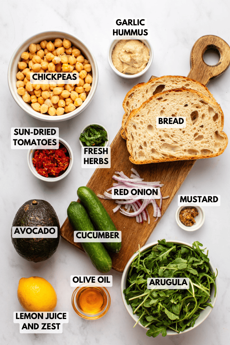 ingredients for a vegan smashed chickpea salad sandwich, including chickpeas, hummus, avocado, cucumber, arugula, bread, herbs, mustard, and sun-dried tomatoes.