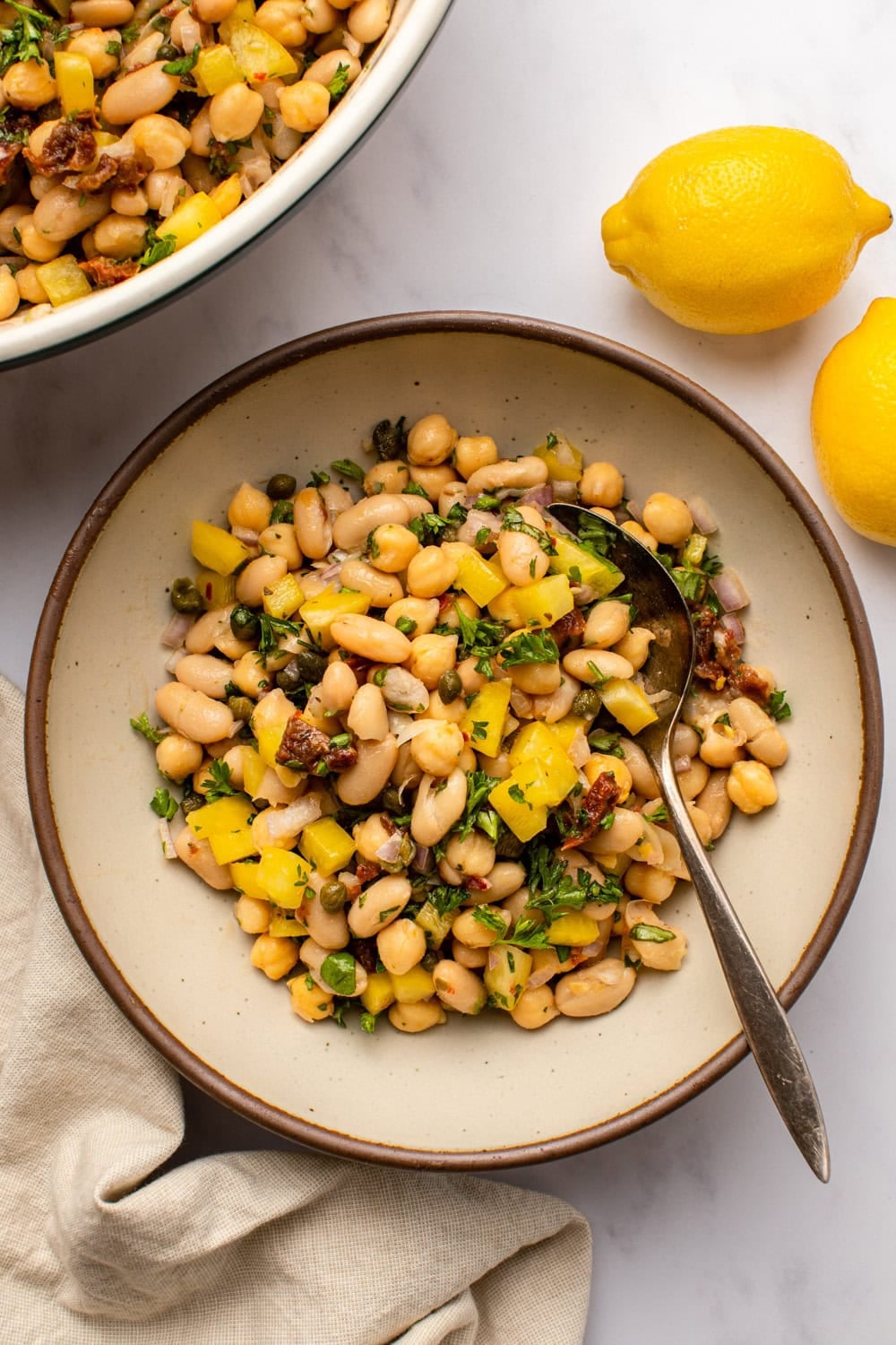a large bowl of lemon caper bean salad with white beans, chickpeas, fresh herbs, yellow bell pepper, capers, and sun-dried tomatoes, tossed together with a light dressing.