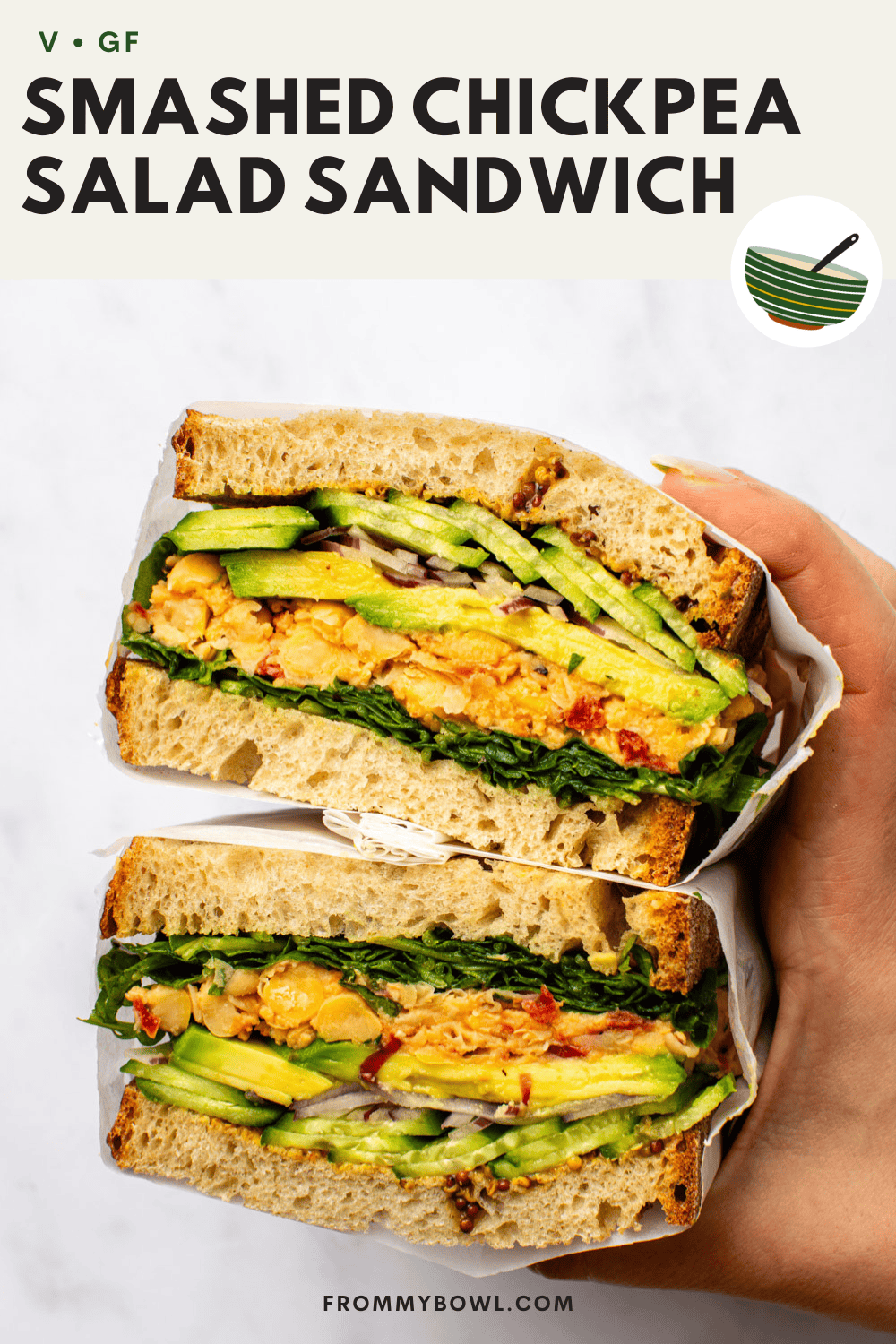 a vegan smashed chickpea salad sandwich layered with avocado, cucumber, red onion, and leafy greens on rustic bread, held in hand.