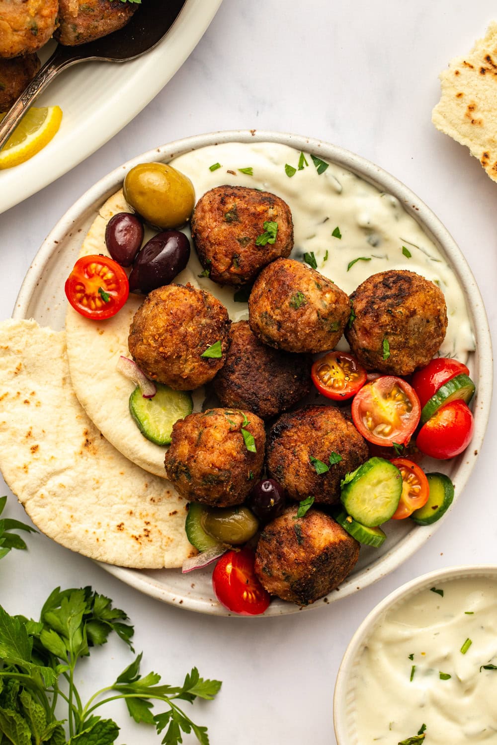 a white ceramic plate sits on a kitchen countertop, holding pita bread topped with vegan Greek-style meatballs, diced tomatoes, cucumber, olives, and a generous drizzle of tzatziki. fresh parsley and a small bowl of additional tzatziki are arranged nearby.