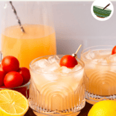 two glasses of sparkling tomato lemonade on a wood cutting board, with jars of lemonade and tomato simple syrup in the background